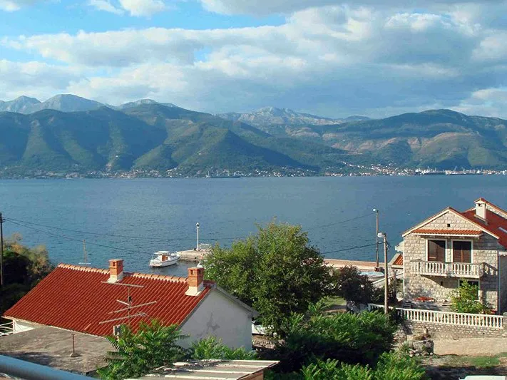 Panoramablick auf das Meer und die Berge vom Haus in Krašići, Montenegro: Steinhäuser mit roten Ziegeldächern und ein Bootssteg.