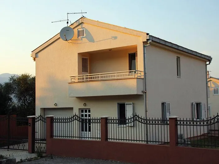 Zweistöckiges Einfamilienhaus mit Balkon und Garage in Krašići, Montenegro, umgeben von einem schmiedeeisernen Zaun vor Bergkulisse.