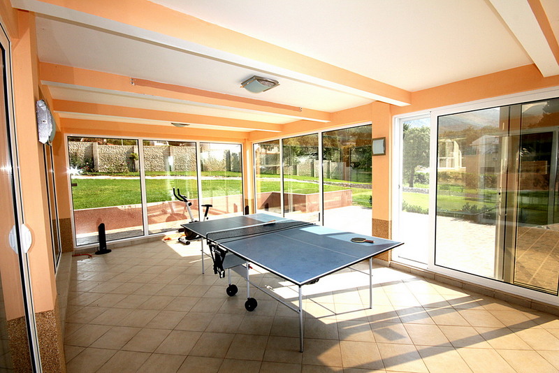 Ein helles Sonnenzimmer mit großen Fenstern, einer Tischtennisplatte und einem Heimtrainer.