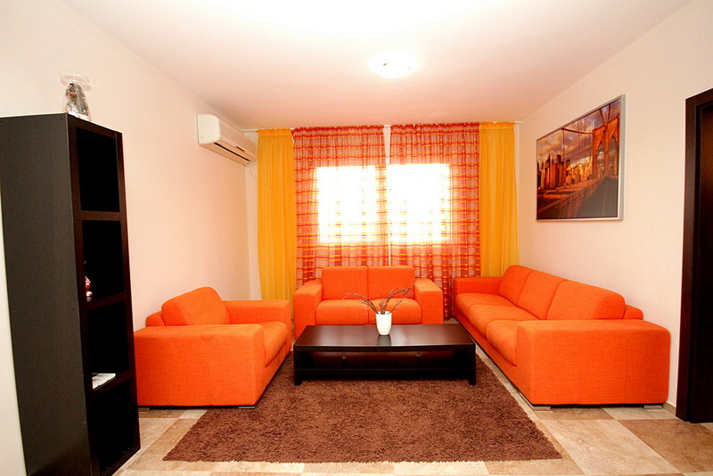 Helles Wohnzimmer mit orangen Sofas, dunklen Holzregalen, Couchtisch und großem Fenster mit orangen Vorhängen.