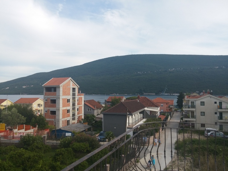 Pogled s balkona na more i planine u Đenovićima, Crna Gora: panorama naselja sa kućama i kovanom ogradom terase.