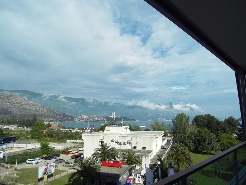 Blick vom Balkon einer Wohnung in Budva, Montenegro: Panorama auf Berge, Meer und die städtische Infrastruktur mit Palmen.