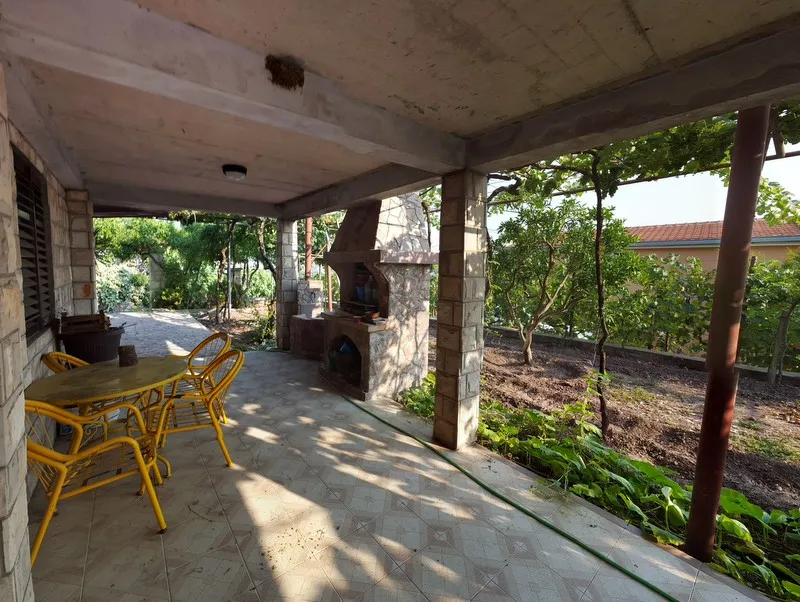 Überdachte Terrasse mit Steingrill und gelben Möbeln in einem Haus in Šušanj, Montenegro, mit Blick auf den grünen Garten und den Weinberg.