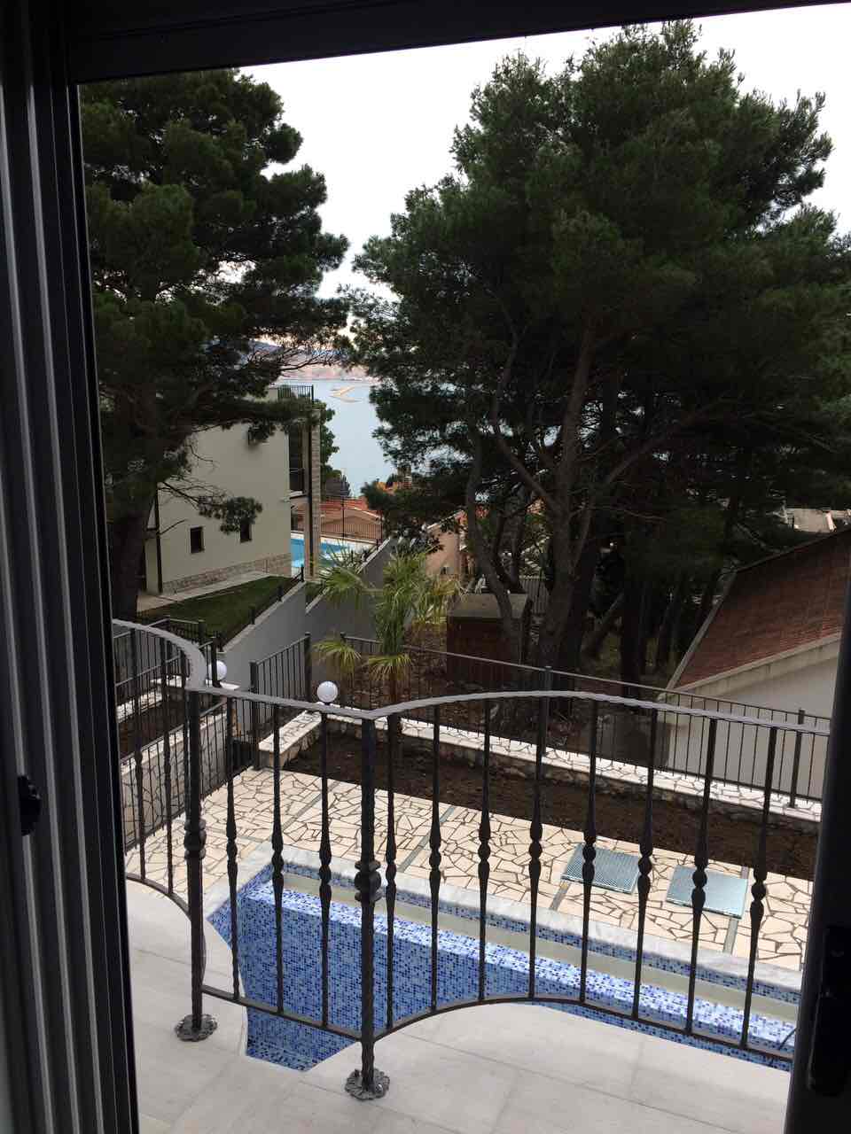 Blick vom Balkon des Hauses in Šušanj, Montenegro: Pool mit Mosaik, Terrasse, Nadelbäume und Meereslandschaft im Hintergrund.