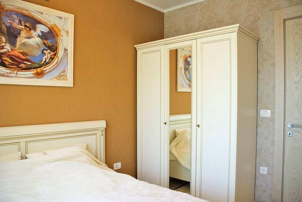 Chambre lumineuse avec mobilier classique et tableau dans un appartement à Kostanica, Monténégro : lit blanc et armoire avec miroir.