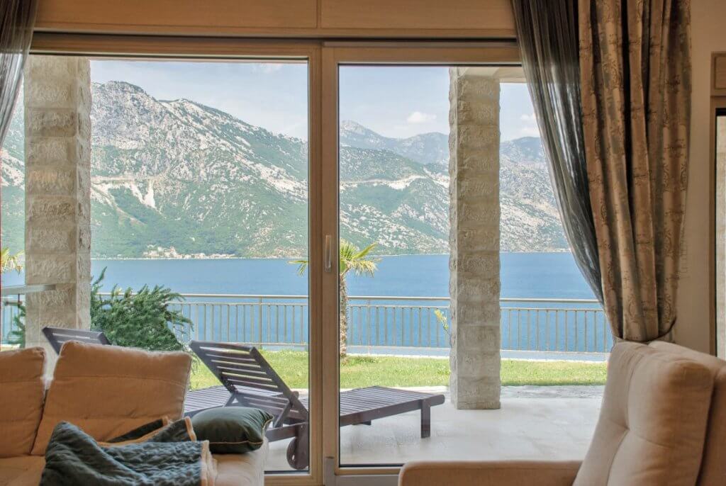 Vue depuis le salon sur la mer et les montagnes dans un appartement à Kotor, Monténégro : baies vitrées panoramiques, terrasse avec chaises longues et colonnes en pierre.