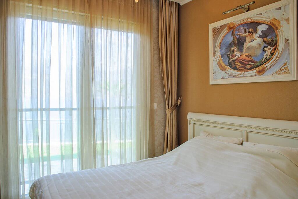 Chambre cosy avec lit blanc et tableau dans un appartement à Kostanica, Monténégro, vue sur la terrasse à travers une baie vitrée panoramique.
