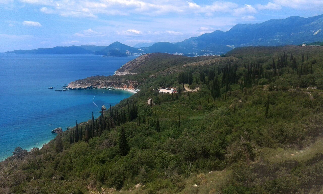 Panoramablick auf die Adria und die Berge von einer Villa in Reževići, Montenegro: grüner Hang, Zypressen und ein Naturstrand.