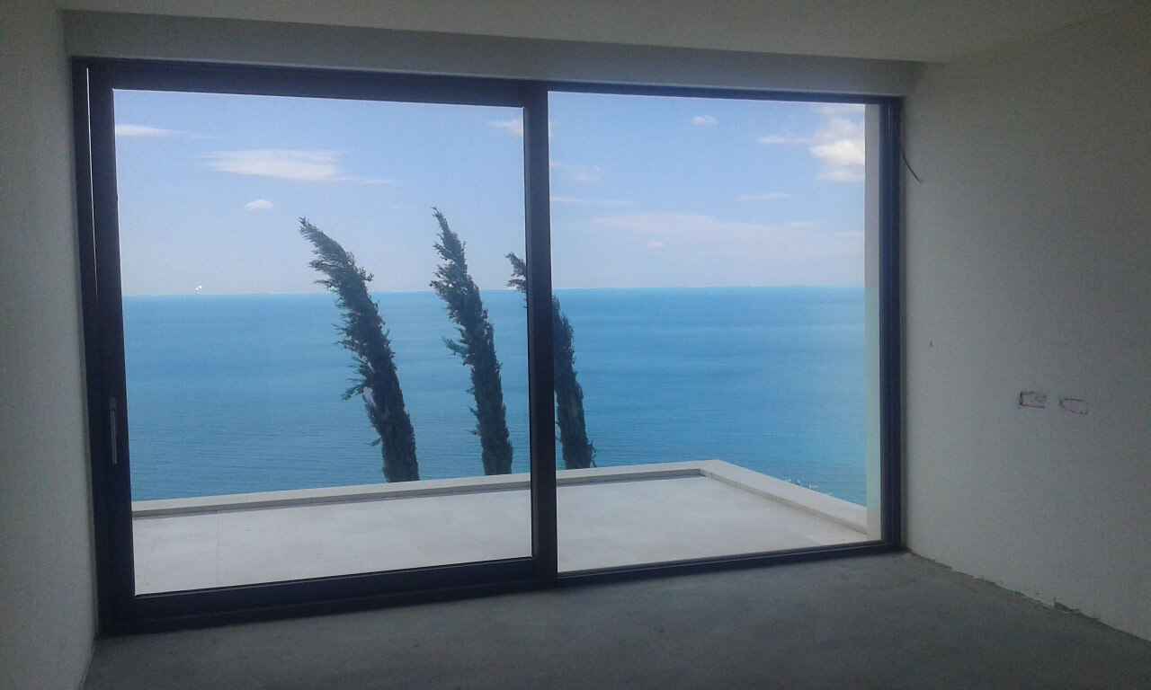 Panoramafenster im Wohnzimmer der Villa in Reževići, Montenegro, mit Blick auf das Meer und die Terrasse mit Zypressen.