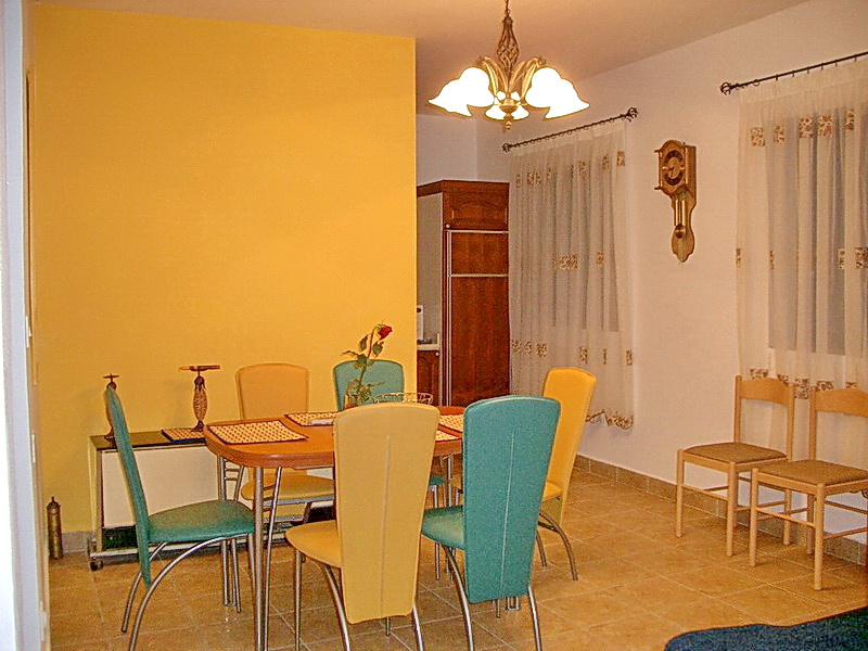 Innenraum des Wohnzimmers mit Essbereich in einem Hotel in Budva, Montenegro: gelbe Wand, Holztisch und bunte Stühle.