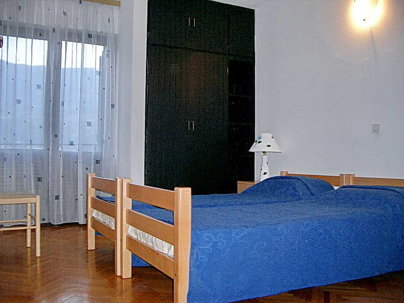 Schlafzimmer mit zwei Betten, dunklem Kleiderschrank und Parkett im Hotel in Budva, Montenegro. Blick auf den Balkon mit transparenten Vorhängen.