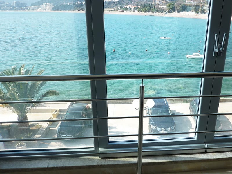 Blick auf das Meer und den Strand vom Balkon der Wohnung in Rafailovići, Montenegro: Panoramafenster, Metallgeländer und Parkplatz.