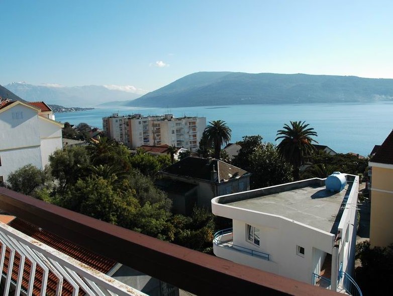 Blick vom Balkon eines Hauses in Herceg Novi, Montenegro: Panorama auf die Bucht von Kotor, Berge, Palmen und städtische Bebauung.