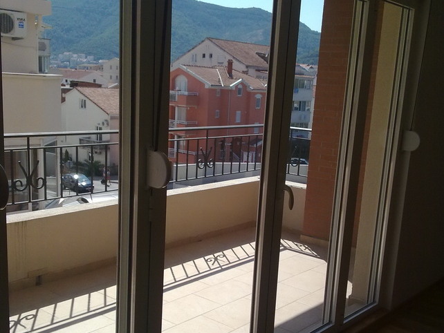 Blick vom Balkon der Wohnung in Budva, Montenegro: Panoramafenster, geflieste Terrasse und Blick auf die Stadthäuser und Berge.