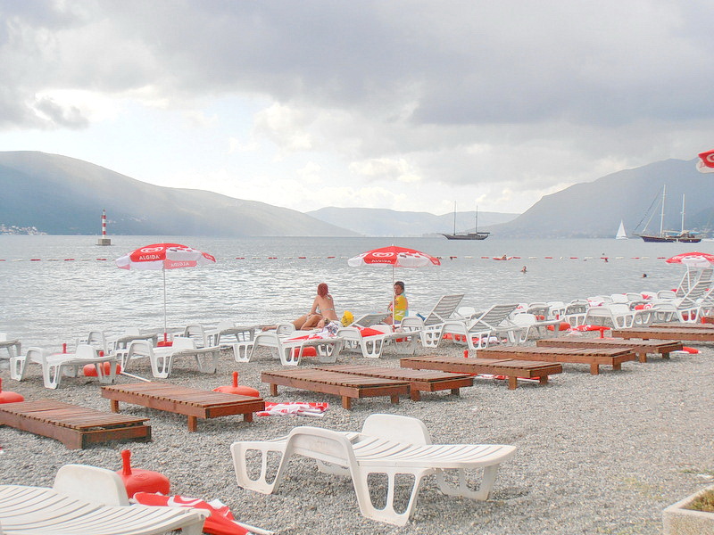 Plaža sa ležaljkama i suncobranima uz more u Tivtu, Crna Gora: pogled na zaliv, planine i jahte pored hotela.