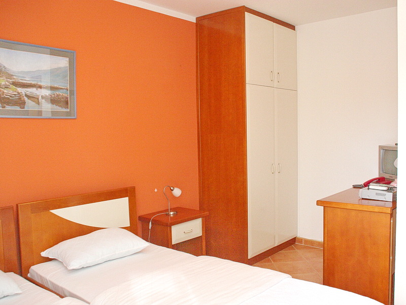 Spavaća soba u hotelu u Tivtu, Crna Gora: dva kreveta, drveni ormar, noćni stočić i narandžasti zid sa slikom.