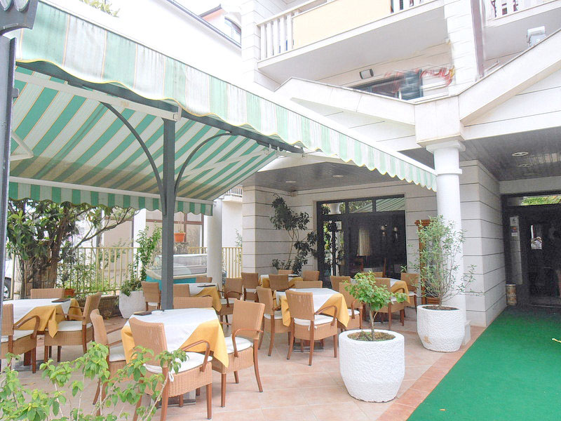 Ugodna letnja terasa hotelskog restorana u Tivtu, Crna Gora, sa stolovima, pletenim stolicama i zelenim nadstrešnicom.
