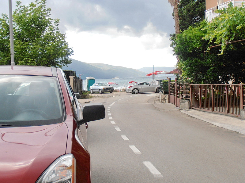 Pogled na put ka moru i plažu pored hotela u Tivtu, Crna Gora, sa parkiranim automobilima i zelenim drvećem.