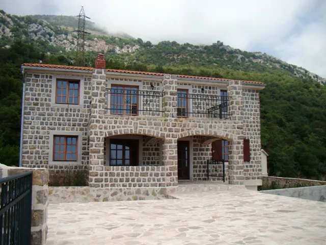 Ein zweistöckiges Steinhaus mit Balkon und gepflastertem Hof vor grünen Bergen.