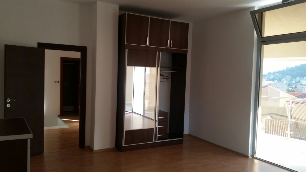 Spavaća soba u stanu u Budvi, Crna Gora: tamni ugradni ormar sa ogledalom, parketni pod i izlaz na balkon s pogledom na grad.