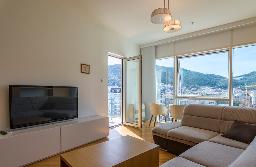 Karadağ, Budva’daki bir dairede modern oturma odası: dağ manzaralı panoramik pencereler, açık renkli mobilyalar ve TV alanı.