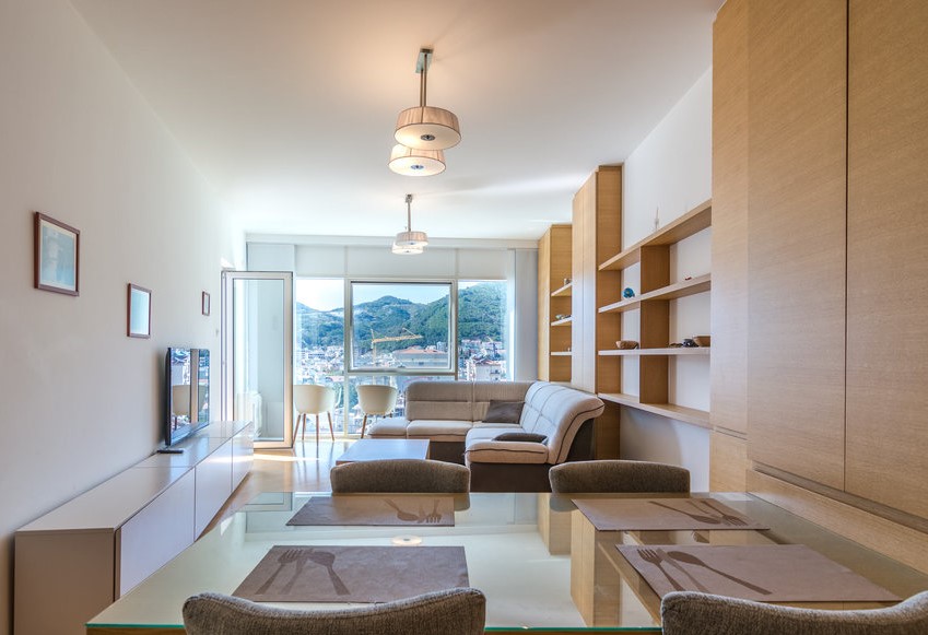 Karadağ, Budva’daki bir dairede yemek alanına sahip modern oturma odası: açık renkli mobilyalar, dağların panoramik manzarası.