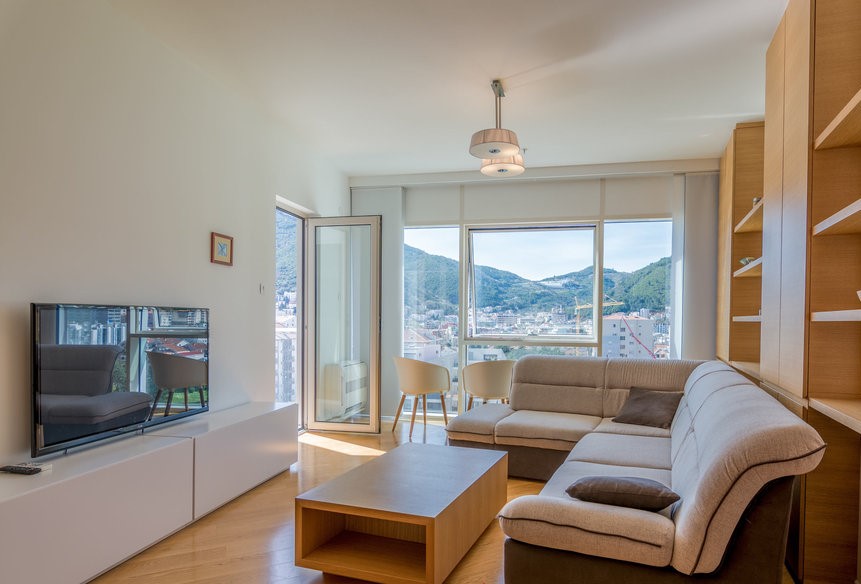 Karadağ, Budva’daki bir dairede modern oturma odası: köşe koltuk, dağlara ve şehre bakan panoramik pencereler.