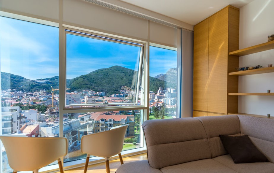Budva, Karadağ’daki dairede oturma odası: dağ manzaralı panoramik pencereler, modern mobilyalar ve aydınlık iç mekân.
