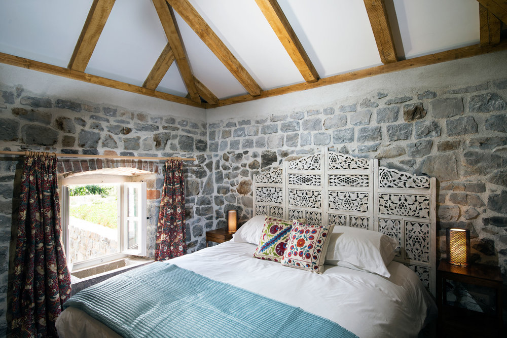 Schlafzimmer in einer Villa in Luštica, Montenegro: Steinwände, Holzbalken, geschnitztes Kopfteil des Bettes und Blick aus dem Fenster.