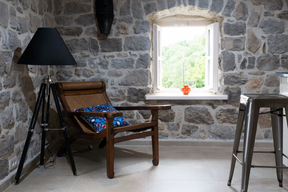 Innenraum des Wohnzimmers in einer Villa in Luštica, Montenegro: Steinwände, Vintage-Sessel, Stehlampe und Blick aus dem Fenster auf den Garten.