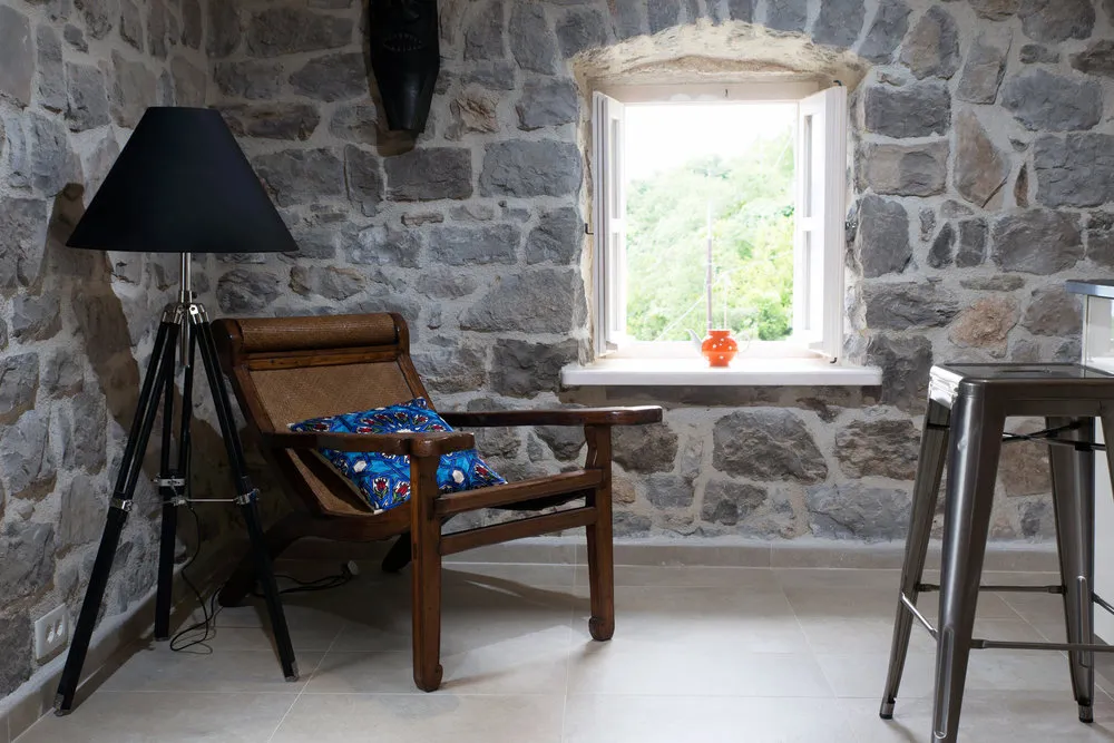 Innenraum des Wohnzimmers in einer Villa in Luštica, Montenegro: Steinwände, Vintage-Sessel, Stehlampe und Blick aus dem Fenster auf den Garten.