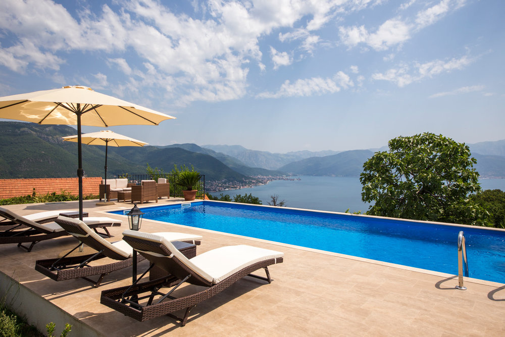 Pool mit Panoramablick auf die Bucht von Kotor und die Berge in einer Villa in Luštica, Montenegro, mit Liegestühlen und Sonnenschirmen.