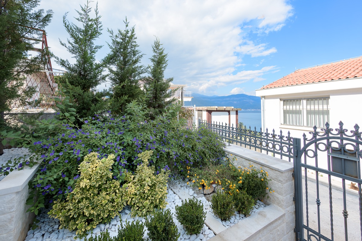 Gepflegter Garten mit blühenden Sträuchern und Meerblick an einer Villa in Krašići, Montenegro, mit schmiedeeisernem Zaun und Terrasse.