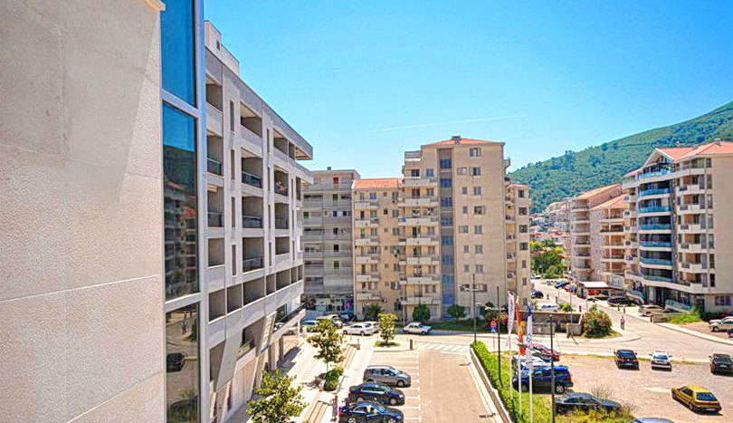 Karadağ’ın Budva şehrinde, güneşli bir günde dağlarla çevrili modern konut kompleksleri ve otopark manzarası.