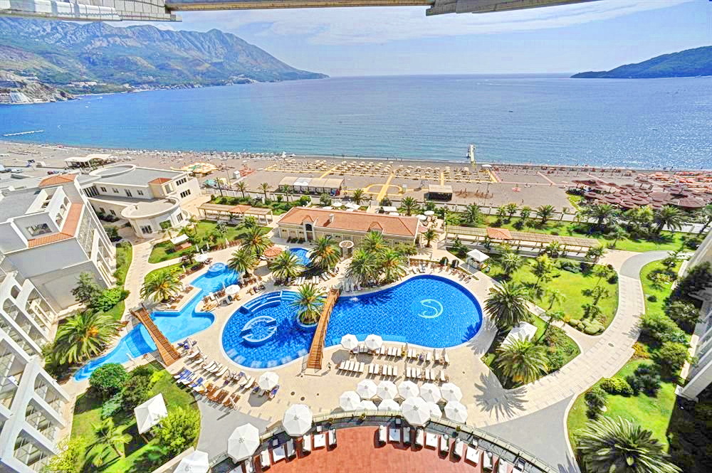 Becici, Karadağ'da balkondan deniz, plaj ve otel havuzuna bakış: palmiyelerle çevrili tatil kompleksinin panoraması.