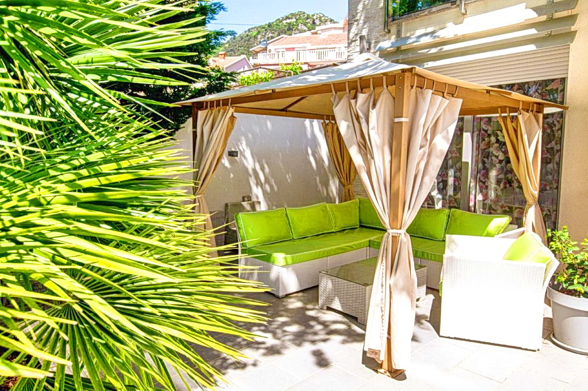 Gemütliche Terrasse mit Pavillon und Rattanmöbeln in einer Villa in Budva, Montenegro, umgeben von Palmen und Grün.