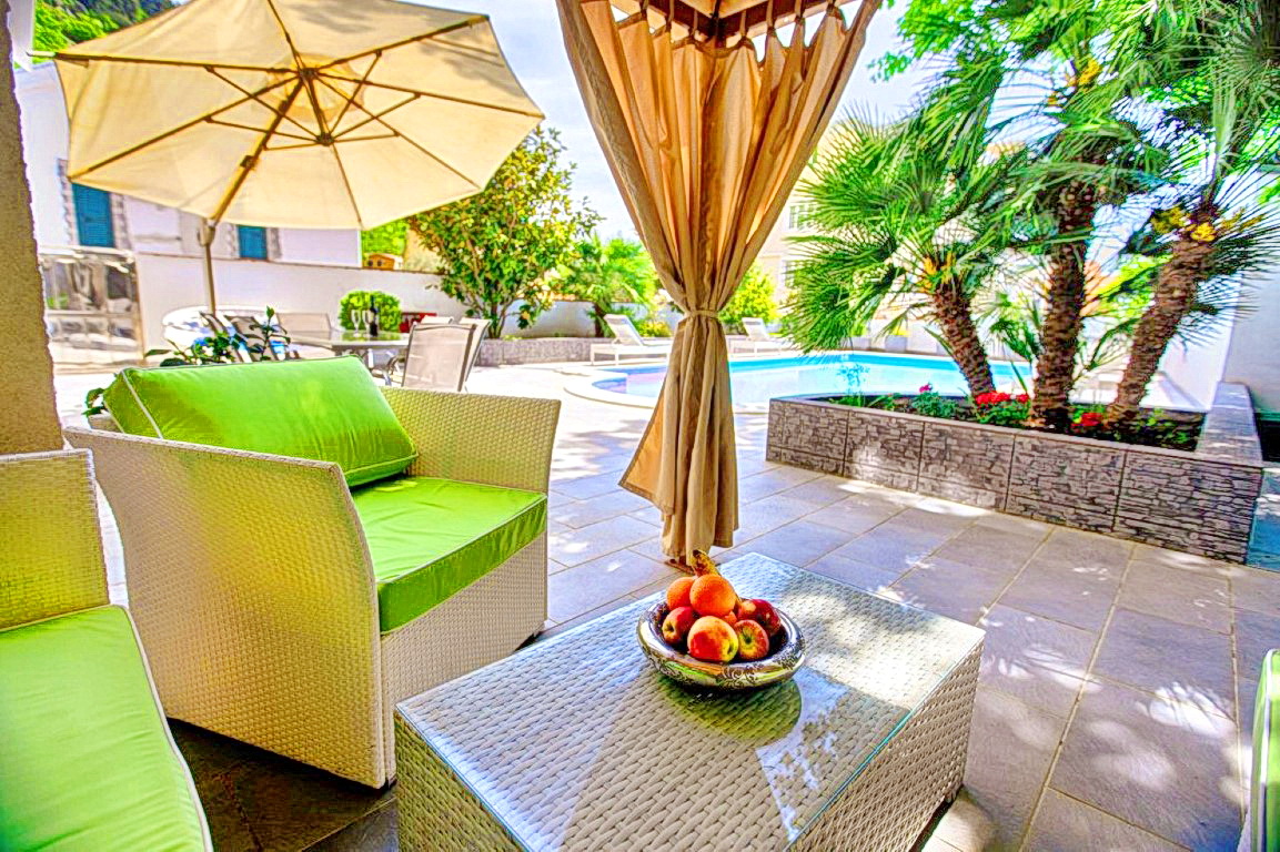 Terrasse der Villa in Budva, Montenegro: geflochtene Möbel, Pool, Palmen und Sonnenschirm in einem gemütlichen Innenhof.