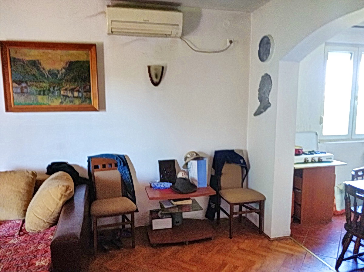 Dnevna soba u kući u Podgorici, Crna Gora: mekana sofa, stolice, klima-uređaj i prolaz do kuhinje sa šporetom.