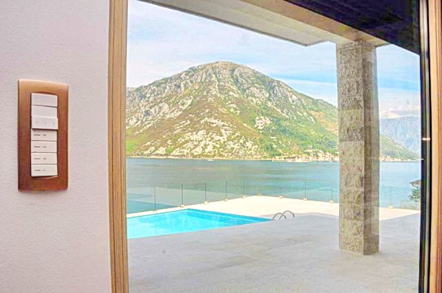 Karadağ, Morinj konut kompleksinde oturma odasından havuza ve Kotor Körfezi’ne manzara: panoramik pencere, taş sütun ve teras.