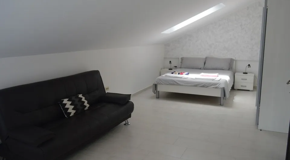 Helles Schlafzimmer im Dachgeschoss einer Villa in Tivat, Montenegro: Doppelbett, schwarzes Sofa und Dachfenster.