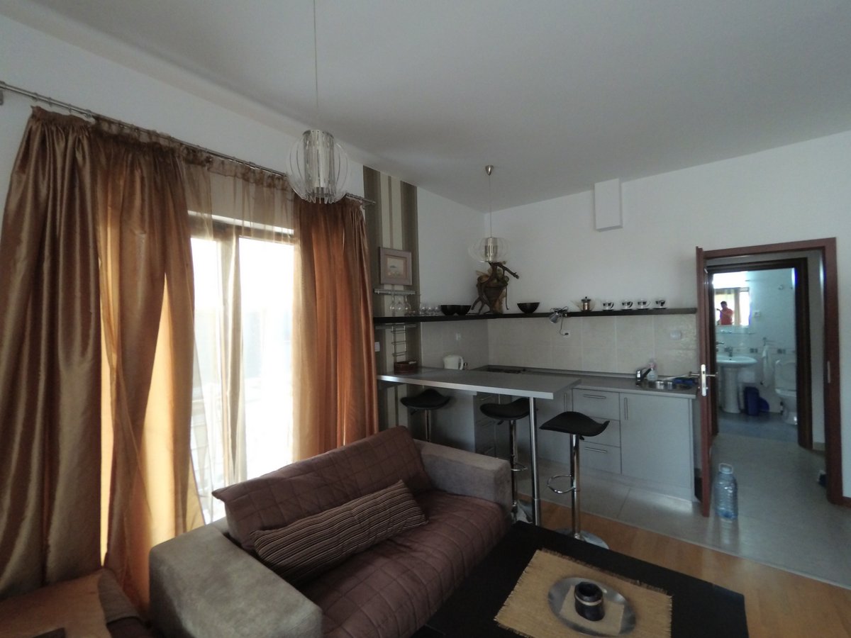 Dnevna soba sa kuhinjom i barskim pultom u stanu u Budvi, Crna Gora: savremeni nameštaj, svetla završna obrada i izlaz na balkon.