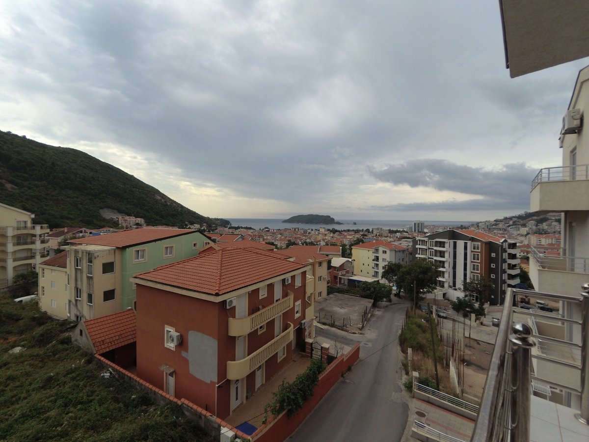 Pogled sa balkona stana u Budvi, Crna Gora: panorama grada, crveni krovovi od crepova, planine i morski zaliv.