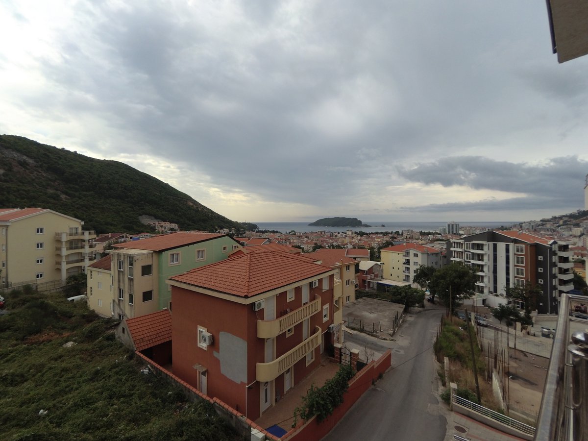 Pogled sa balkona stana u Budvi, Crna Gora: panorama grada, stambene zgrade sa crvenim krovovima i more na horizontu.