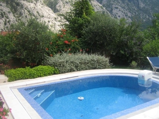 Open-air swimming pool with blue tiles at a villa in Orahovac, Montenegro, against a backdrop of mountains and a green garden with a sun lounger.