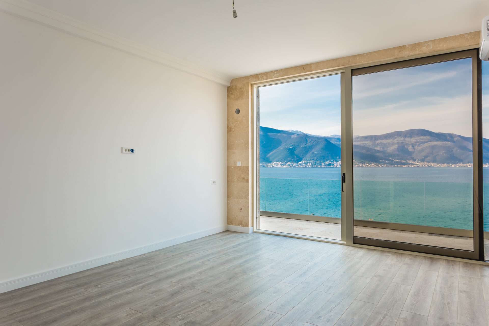 An empty room with a large glass sliding door offering a view of the sea and mountains.