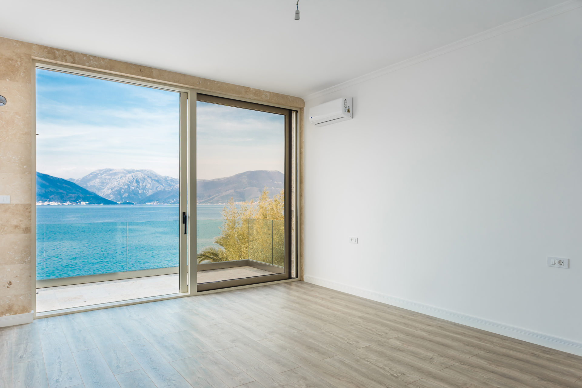 An empty room with a large sliding glass door overlooking the sea and mountains, light wood-look flooring, and white walls.