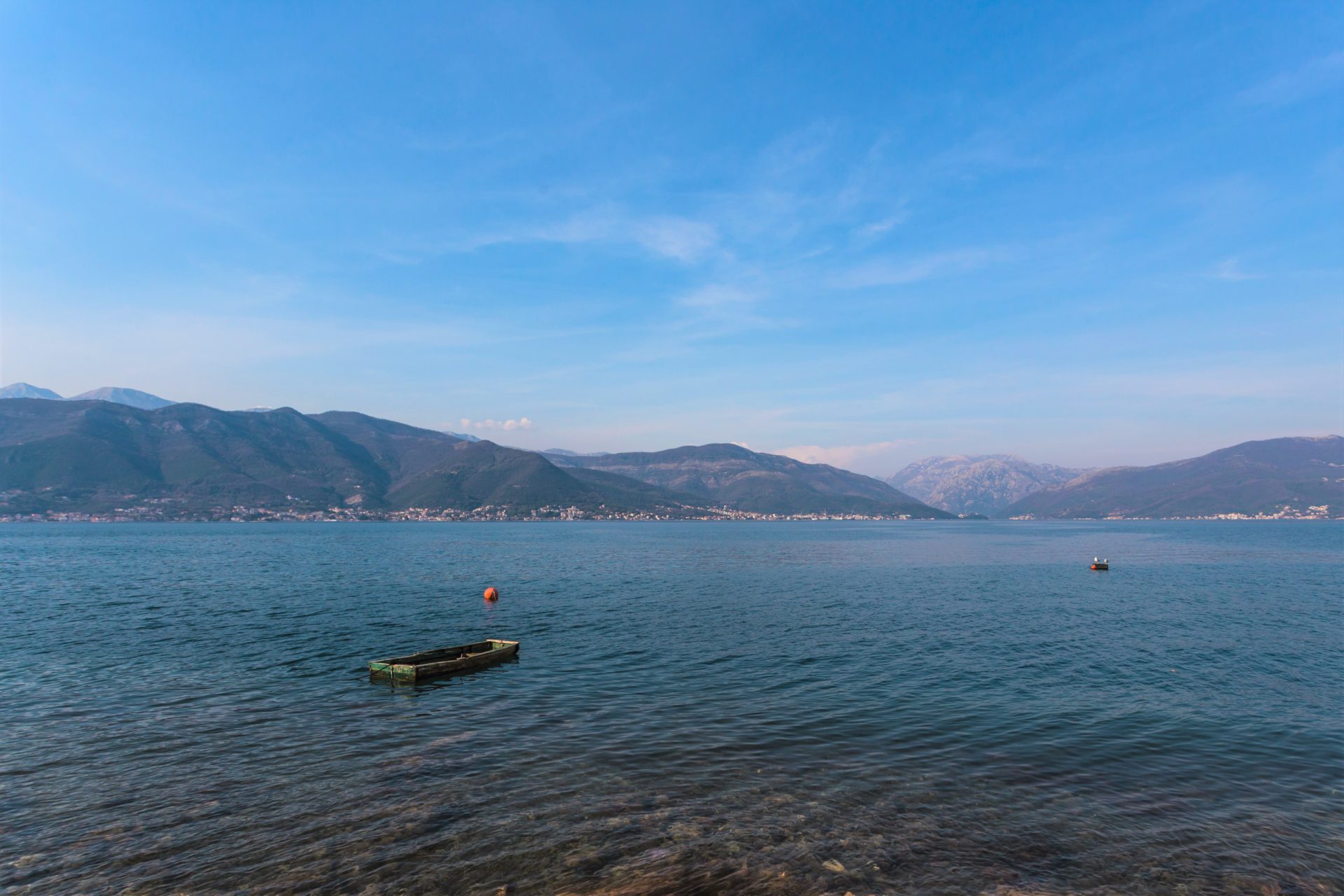 A wide view of a calm blue sea with a small wooden boat, mountains in the distance, and a clear blue sky.