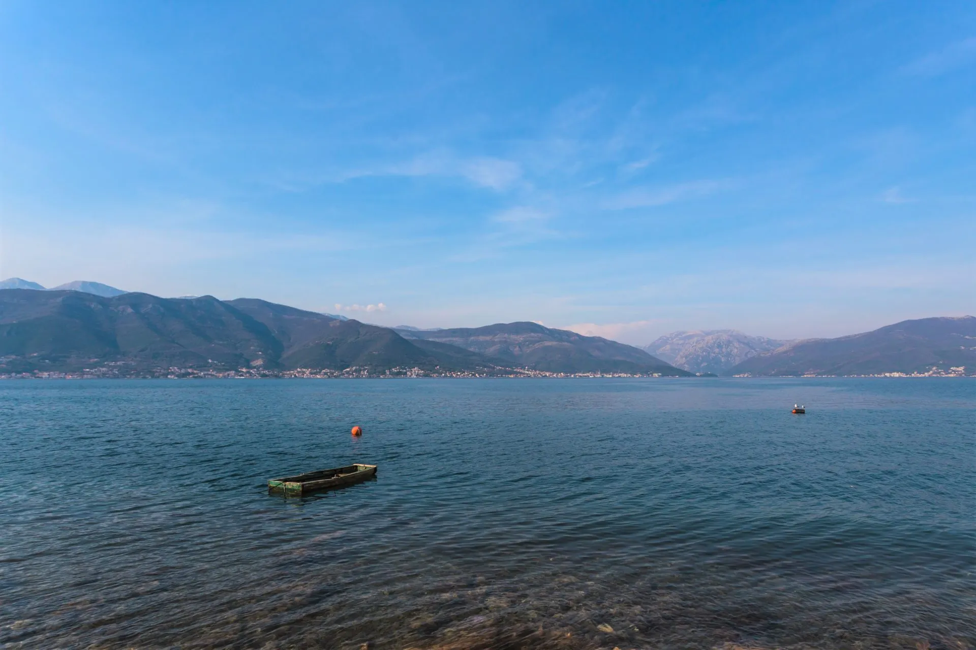Weiter Blick auf ein ruhiges, blaues Meer mit einem kleinen Holzboot, Bergen in der Ferne und klarem blauen Himmel.