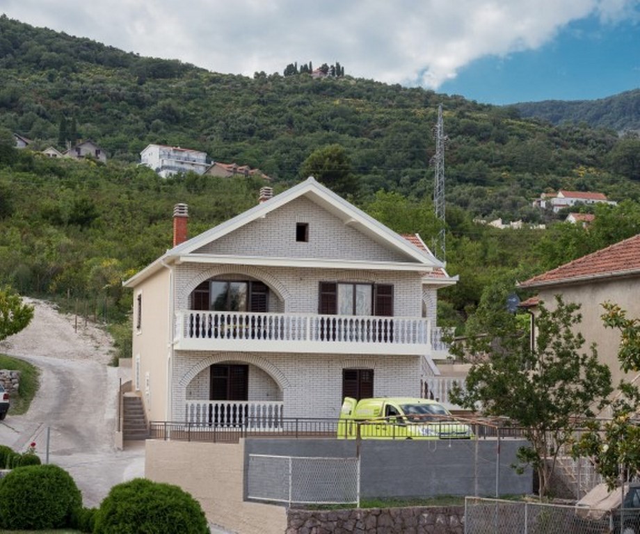 Zweistöckiges Haus aus hellem Ziegelstein mit bogenförmigen Balkonen in der Stadt Bijela, Montenegro, vor dem Hintergrund grüner Berge.