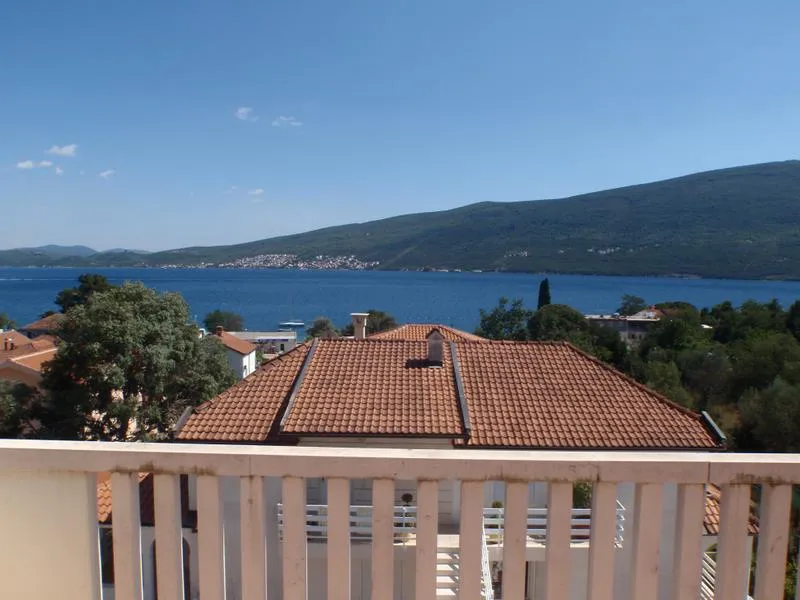 Pogled sa balkona kuće u Baošićima, Crna Gora: panorama mora, planina i krovova od crepova tokom sunčanog dana.